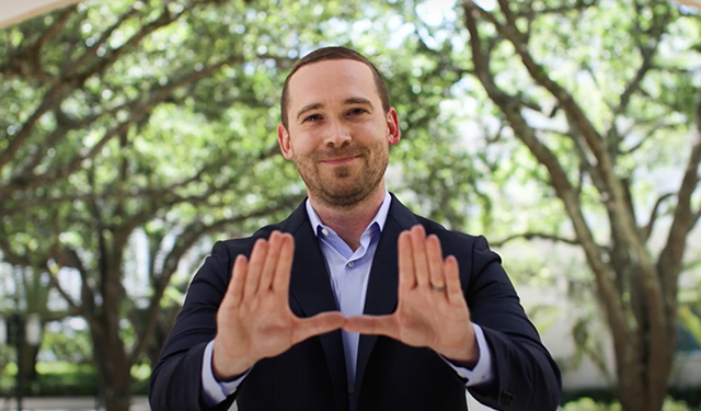 A MAIA alumni makes the U sign with his hands, smiling at the camera. There is dappled light behind him from the canopies of the UM campus. 