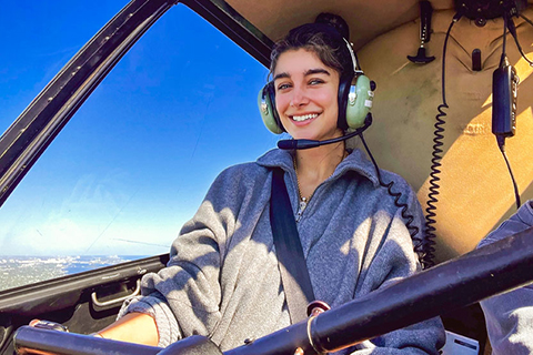 This is a photo of a University of Miami alumnae. She is sitting inside of a helicopter. She is smiling at the camera and wearing a pair of aviator headphones.. 